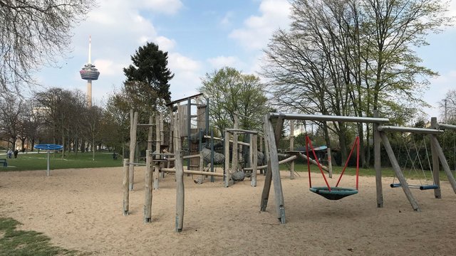 Ein Spielplatz mit vielen Holzelementen ist zu sehen. Im Hintergrund ist der Colonius.