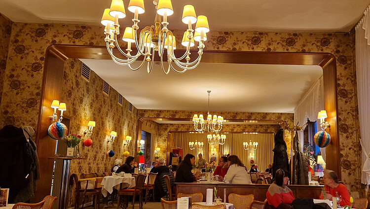 Innenraum des Café Wahlen mit Kronleuchtern, floraler Tapete, Rattanstühlen und Gästen an weißgedeckten Tischen