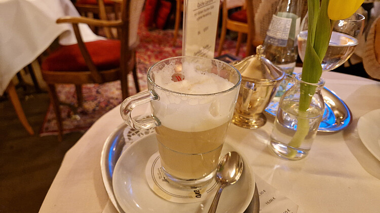 Milchkaffee in klassischer Glastasse mit Milchschaum serviert auf weißer Untertasse mit Löffel